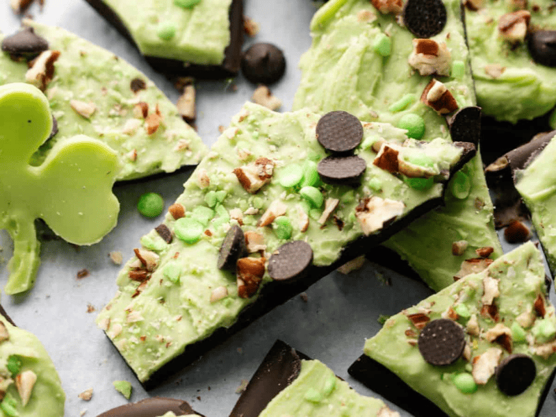 Pieces of green and dark chocolate bark topped with nuts and chocolate chips are scattered on a surface. A shamrock-shaped green chocolate sits on the left side.