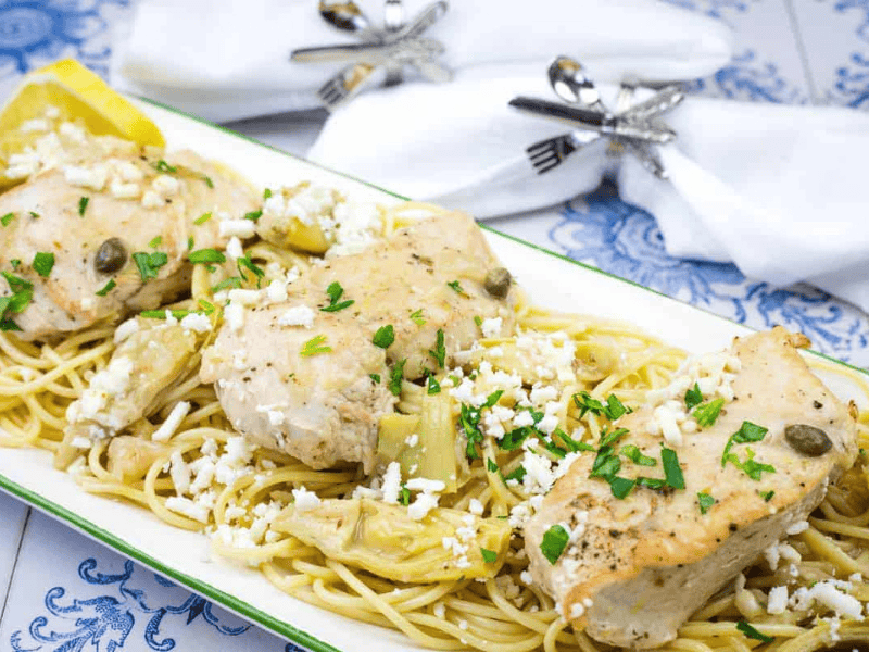 A dish of pasta topped with Skillet Chicken with Artichokes, Lemon, and Feta garnished with parsley.