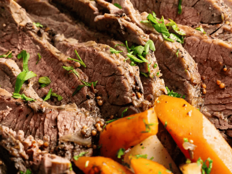 Sliced cooked beef garnished with chopped herbs, served with cooked carrots and potatoes.