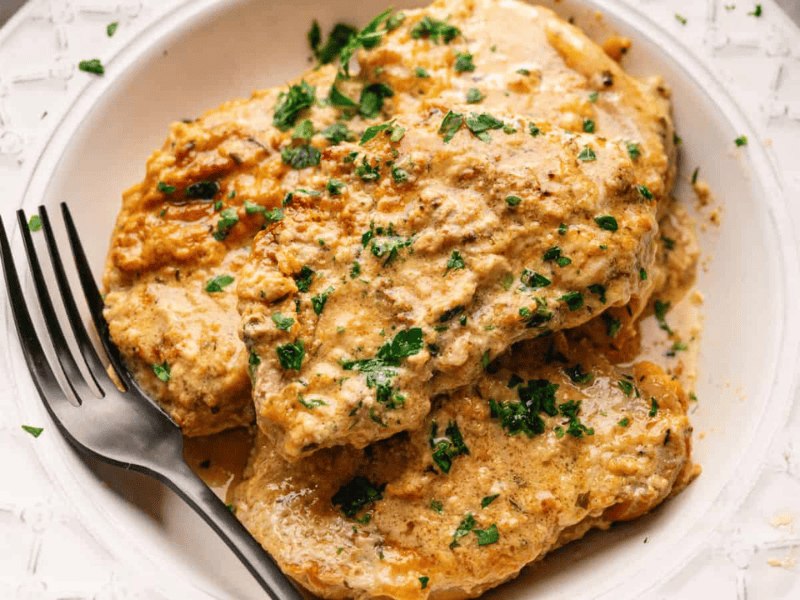 Chicken breasts coated in a creamy, herb sauce are served on a white plate with a fork, garnished with chopped parsley.