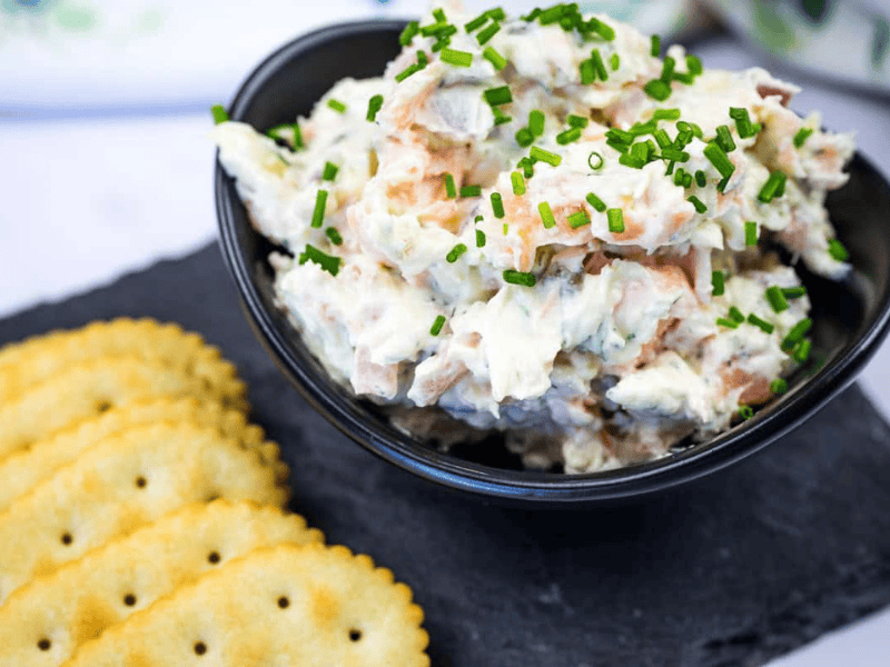 Bowl of smoked salmon spread garnished with chopped chives next to crackers on a slate platter.