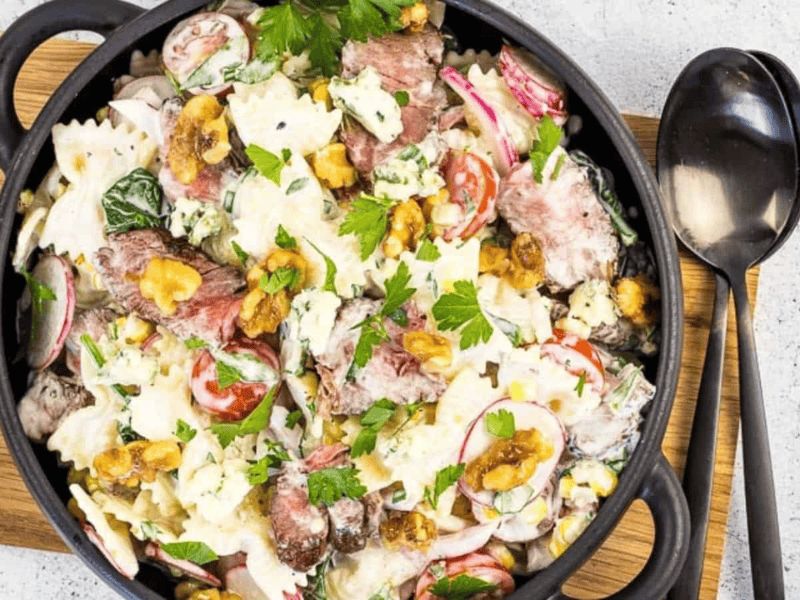 A black bowl filled with a pasta salad containing farfalle, steak slices, radishes, cherry tomatoes, lettuce, nuts, and crumbled blue cheese, garnished with parsley, on a wooden board next to a spoon.