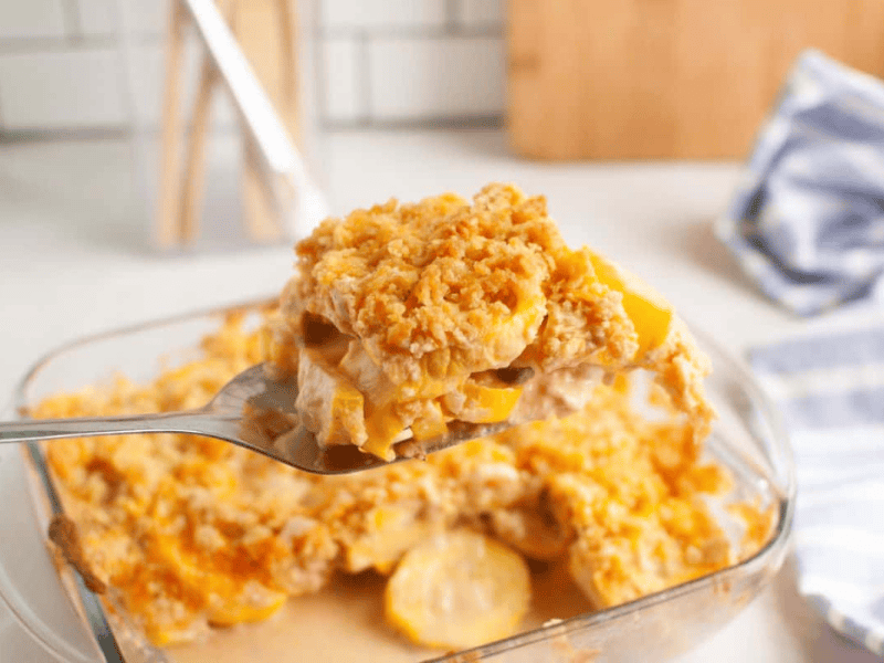 A spoon holds a serving of yellow squash casserole with a crumbly topping above a glass baking dish on a kitchen counter.