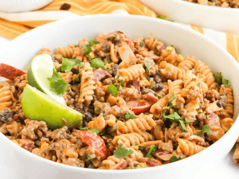 A bowl of fusilli pasta mixed with ground beef, black beans, chopped tomatoes, and herbs, garnished with lime wedges.