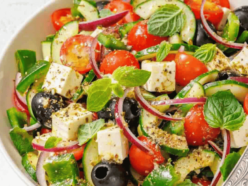A bowl of salad with tomatoes, cucumbers, olives, red onion, feta cheese cubes, and fresh mint leaves, seasoned with herbs.