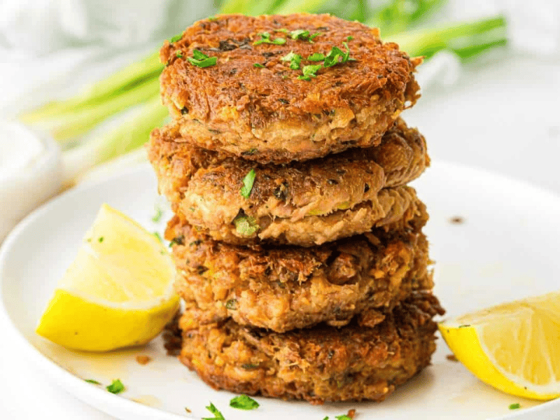 A stack of four golden-brown tuna patties garnished with chopped herbs, served on a white plate with two lemon wedges.
