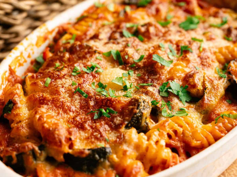 Baked pasta casserole with rotini noodles, tomato sauce, melted cheese, and chopped parsley in a white dish.
