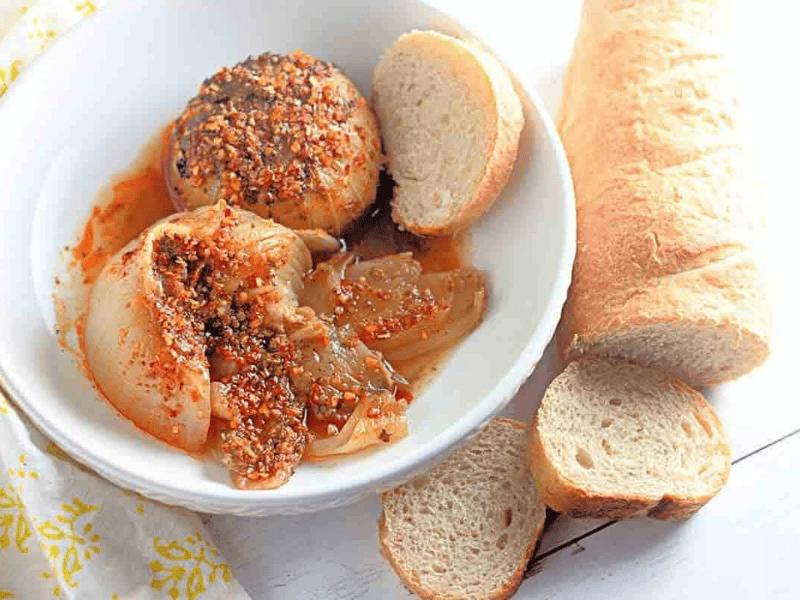 A bowl of cooked onions topped with spices and oil, served with sliced baguette on a white surface.