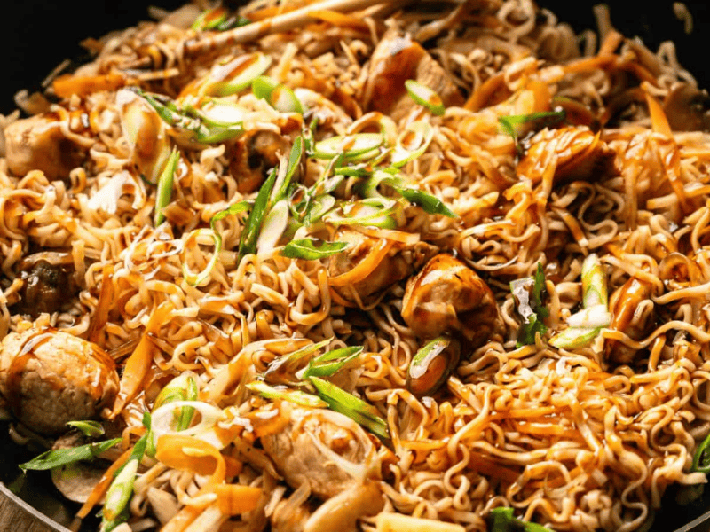 A close-up of cooked stir-fried noodles with pieces of chicken, sliced carrots, green onions, and a savory sauce in a black pan.