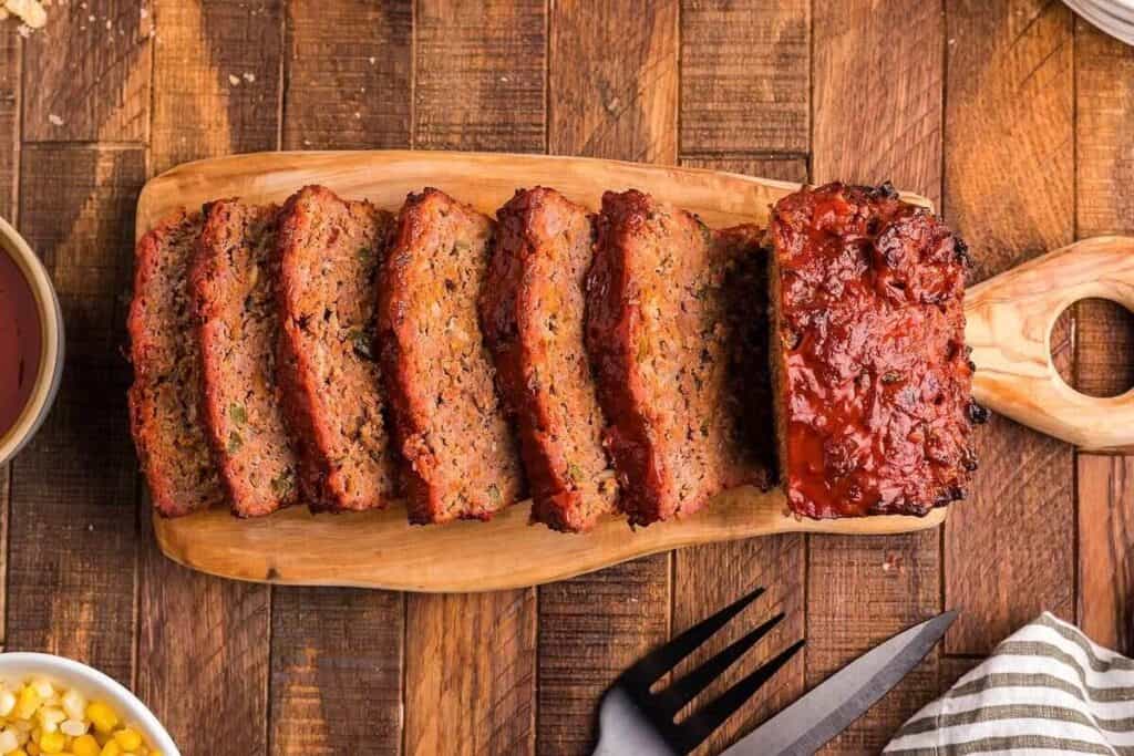 Sliced meatloaf with a tomato glaze sits on a wooden cutting board, surrounded by utensils on a wooden table.