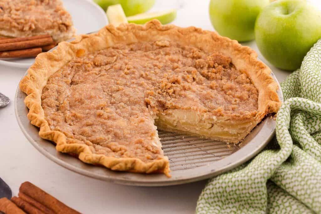 A Dutch apple pie in a metal pie pan has a crumb topping and a missing slice, with green apples, cinnamon sticks, and a green patterned cloth nearby.