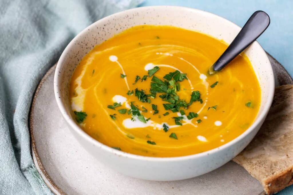 A bowl of creamy orange soup, garnished with a swirl of cream and chopped parsley, sits on a plate next to a piece of bread.