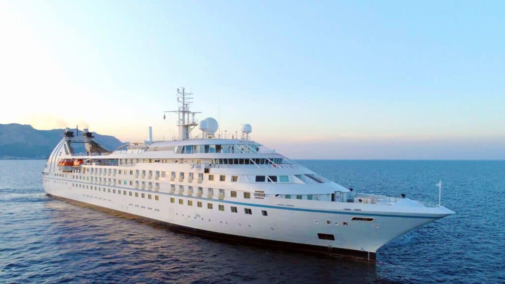 Large white cruise ship sailing on calm blue sea with mountains and a clear sky in the background.