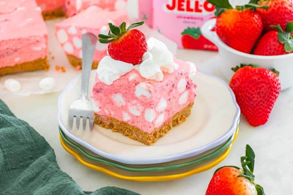 A slice of pink strawberry dessert with marshmallows sits on stacked plates, topped with whipped cream and a strawberry, with fresh strawberries and a Jell-O box in the background.