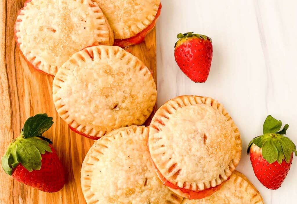 Six round hand pies with golden-brown crusts are arranged on a wooden board next to three whole strawberries on a white surface.
