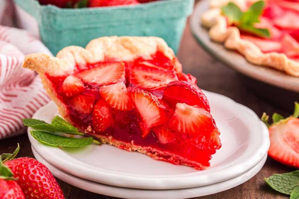 A slice of strawberry pie with a flaky crust, topped with fresh sliced strawberries and red glaze, served on a white plate with mint leaves.