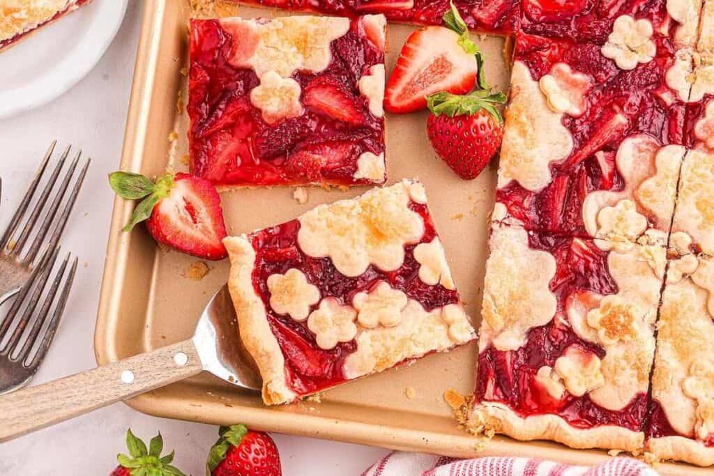 A baking sheet with strawberry slab pie, some pieces cut, decorated with flower-shaped crust, fresh strawberries, and serving utensils nearby.