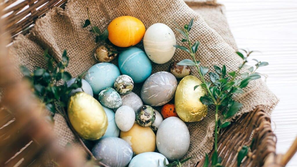 A wicker basket lined with burlap holds various colored and decorated eggs, including blue, gold, yellow, and speckled eggs, with green sprigs around them.