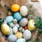 A wicker basket lined with burlap holds various colored and decorated eggs, including blue, gold, yellow, and speckled eggs, with green sprigs around them.