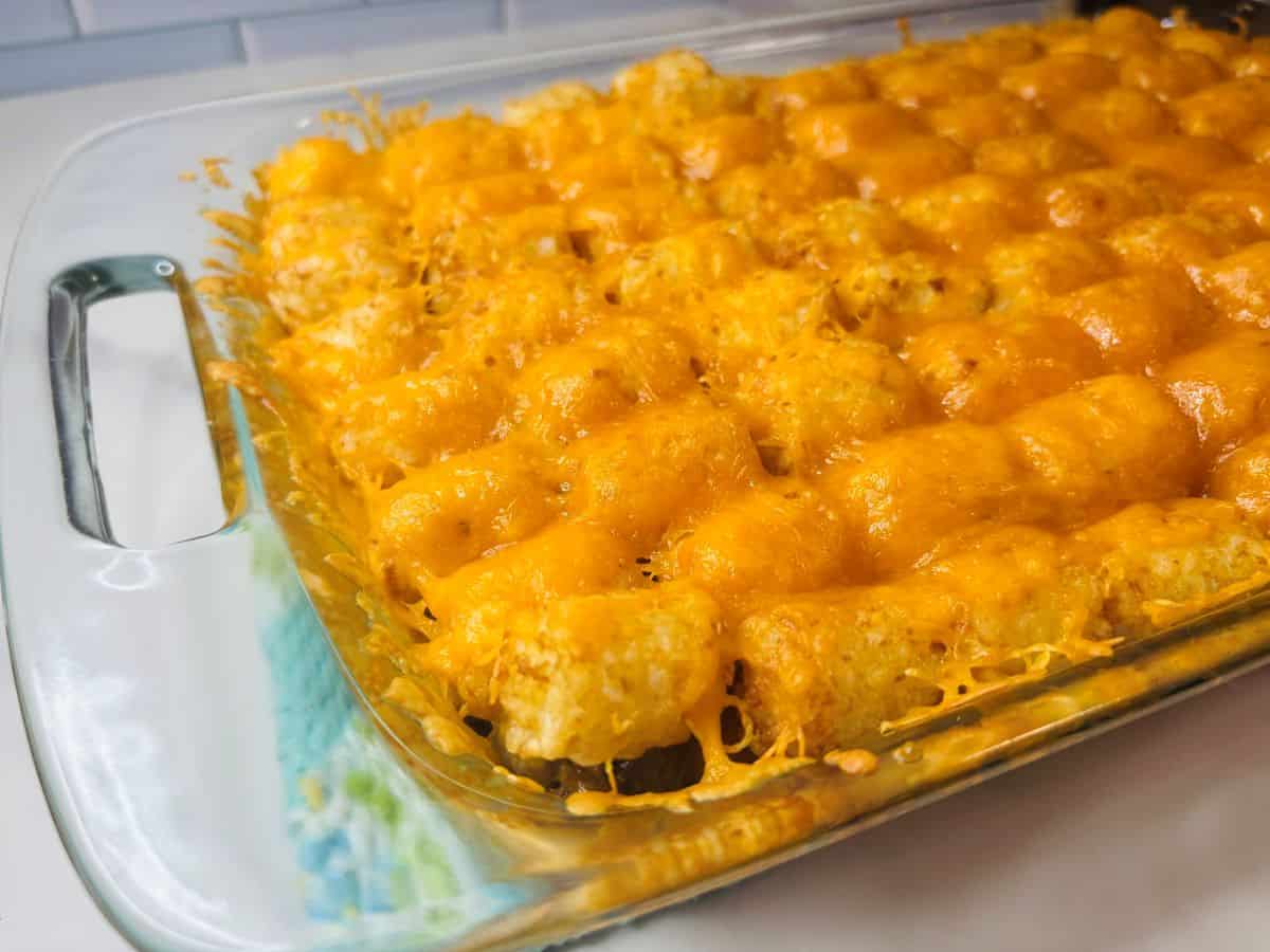 A spatula lifts a portion of a baked casserole featuring tater tots, ground beef, and melted cheddar cheese from a glass dish.