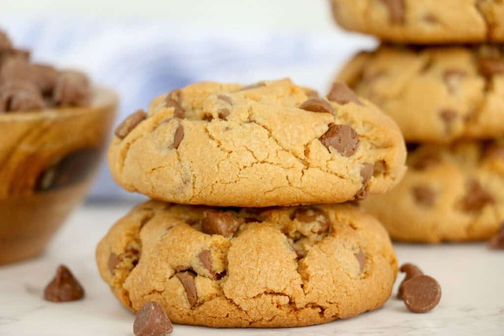 Two chocolate chip cookies stacked on a marble surface with more cookies and a bowl of chocolate chips in the background.