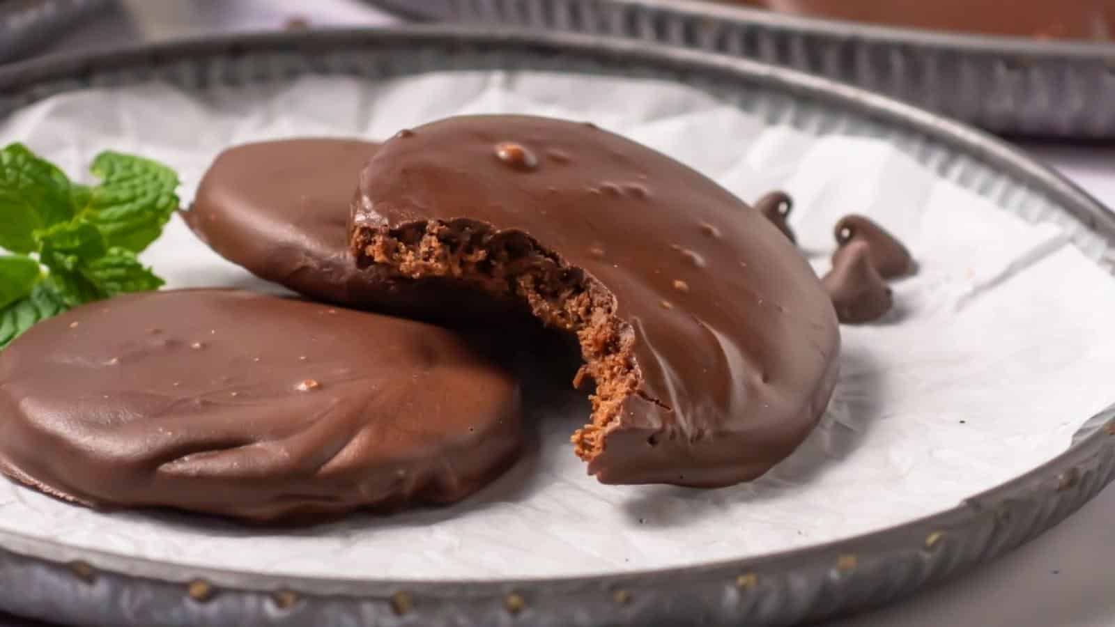 Two copycat thin mint cookies on a plate, one with a bite taken out, with a mint leaf garnish beside them.