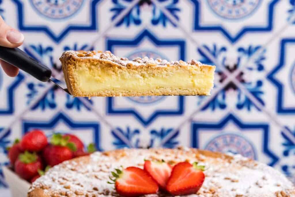 A hand holds a slice of custard tart topped with powdered sugar in front of a blue patterned tile background, with strawberries and the rest of the tart below.