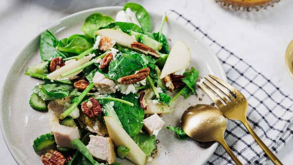 A plate of salad with spinach, pecans, pear slices, cucumber, and chunks of chicken, served with a fork and spoon on the side.
