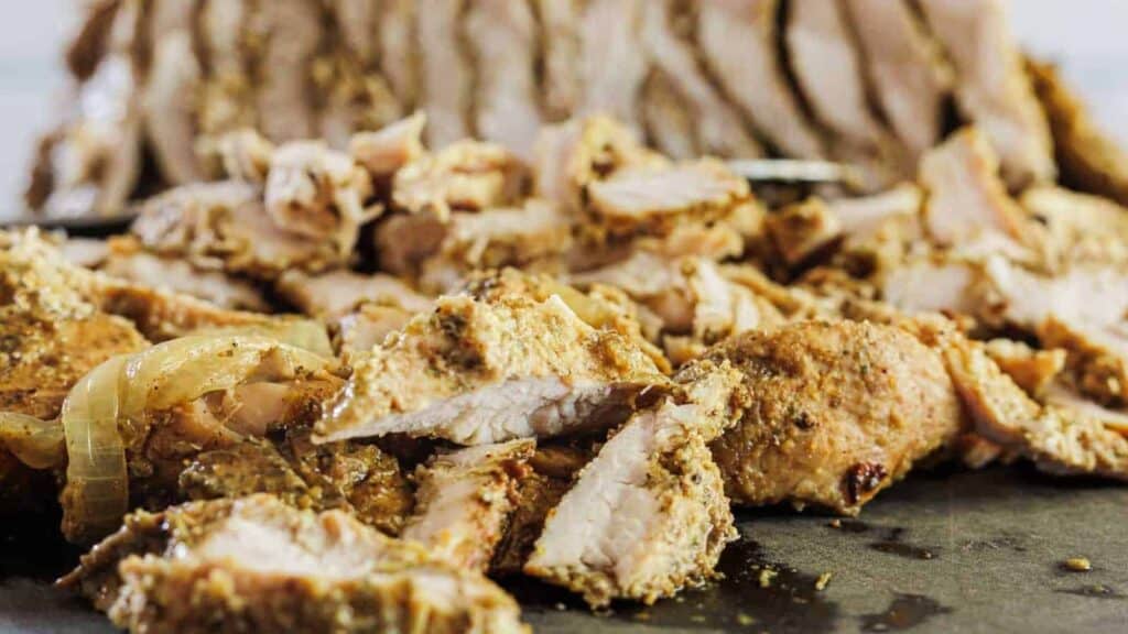 Sliced seasoned cooked pork with visible spices and onions on a dark surface, perfect for steady budget dinners, with more pork in the background.