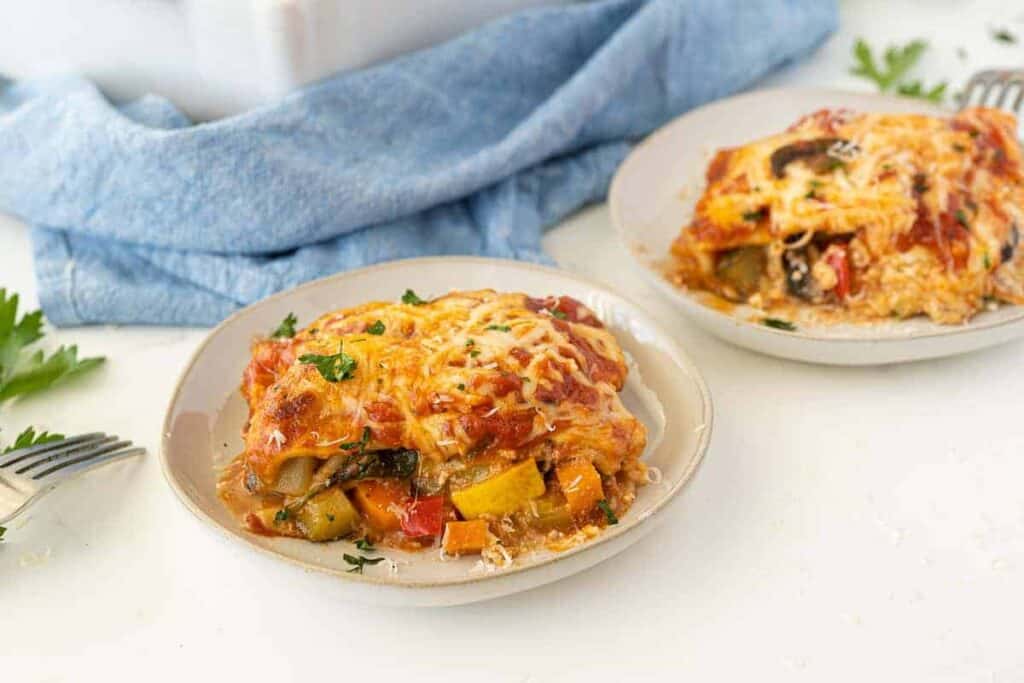 Two plates of vegetable lasagna topped with melted cheese, set on a white surface with a blue cloth and parsley garnish nearby.