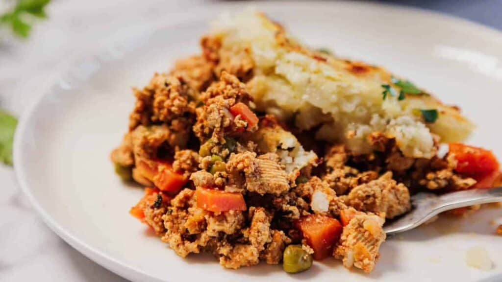A serving of shepherd's pie with ground meat, peas, carrots, and a layer of mashed potatoes on a white plate.