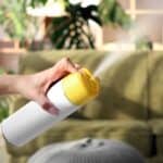 A person sprays an aerosol can with a yellow nozzle in a living room, embracing spring cleaning season amidst a green sofa and lush indoor plants in the background.