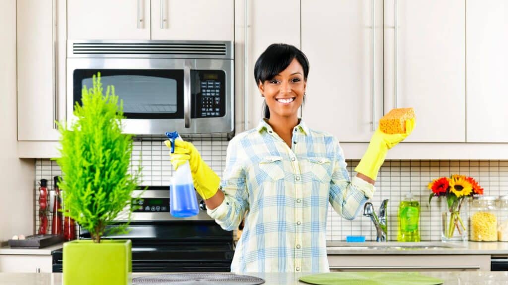 Woman wearing rubber gloves stands in a kitchen holding a sponge and spray bottle, smiling at the camera; kitchen appliances and plants are visible in the background.