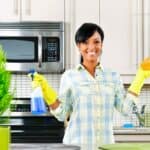 Woman wearing rubber gloves stands in a kitchen holding a sponge and spray bottle, smiling at the camera; kitchen appliances and plants are visible in the background.