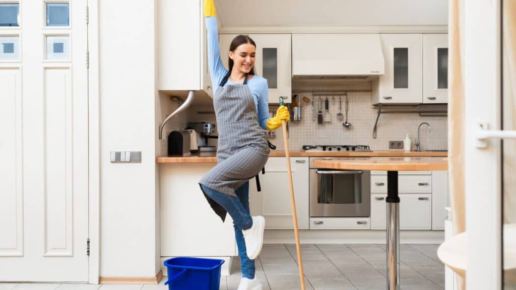 A woman wearing cleaning gloves and an apron poses cheerfully with a mop in a modern kitchen; a blue bucket is on the floor nearby.