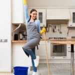 A woman wearing cleaning gloves and an apron poses cheerfully with a mop in a modern kitchen; a blue bucket is on the floor nearby.