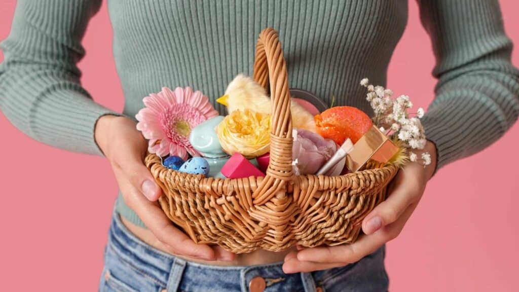 A person holding a wicker basket filled with assorted items like flowers, eggs, and small gifts against a pink background&mdash;perfect inspiration for Easter baskets for adults.