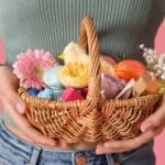 A person holding a wicker basket filled with assorted items like flowers, eggs, and small gifts against a pink background&mdash;perfect inspiration for Easter baskets for adults.