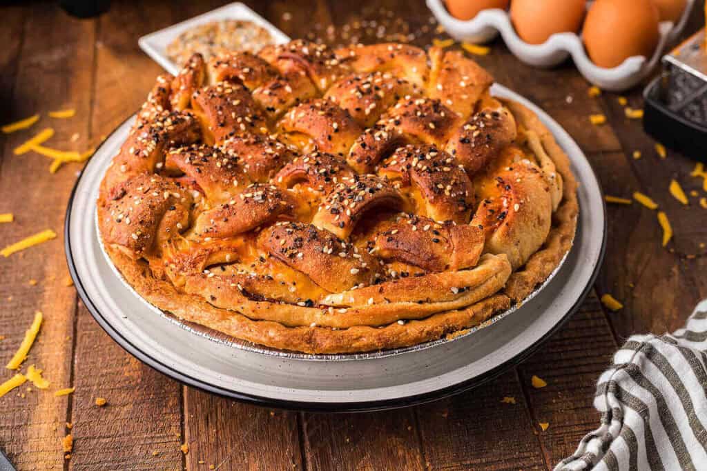 A baked savory pie with a golden, lattice-style crust topped with seeds, sitting on a wooden table near eggs, grated cheese, and a striped cloth.