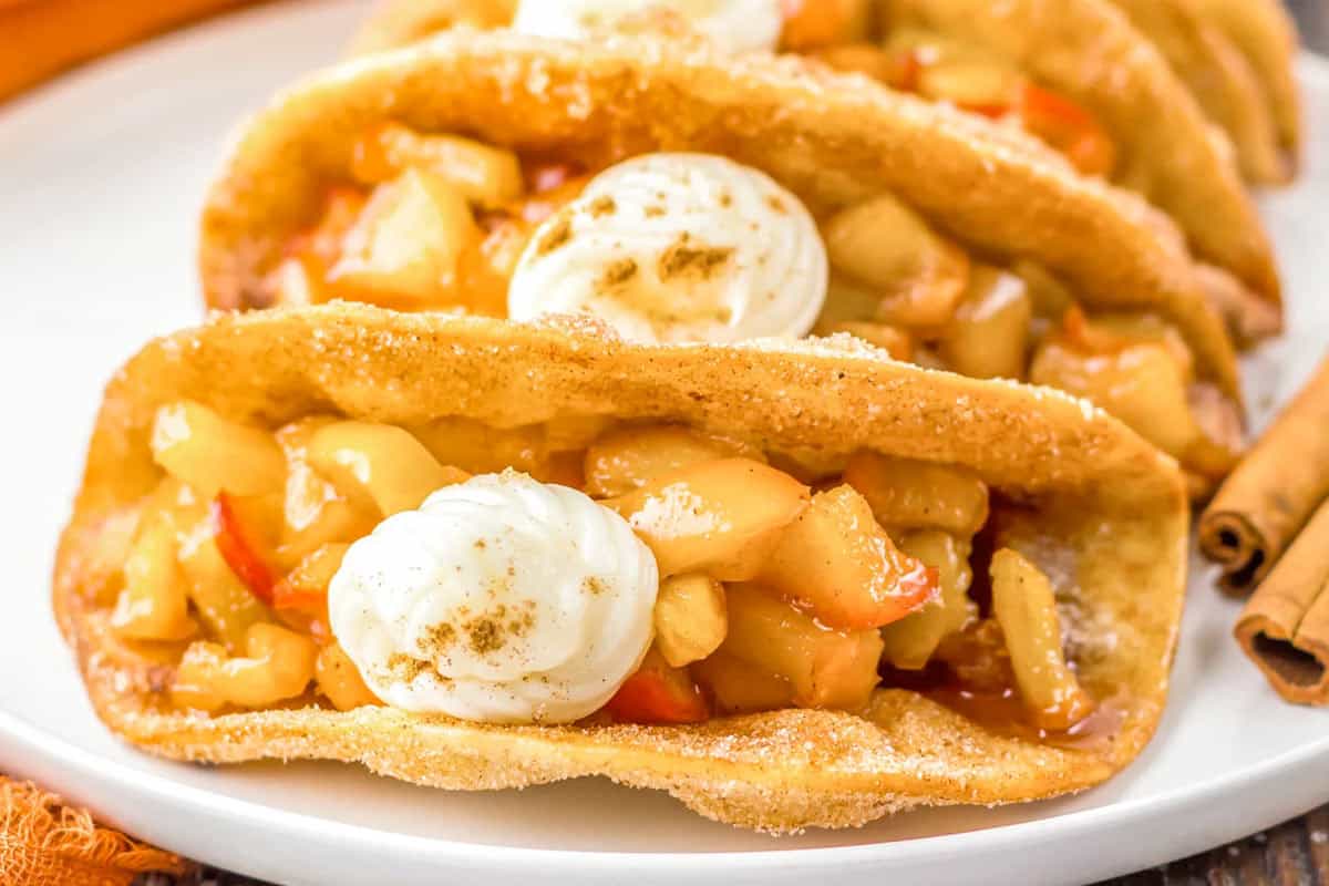 A close-up of dessert tacos on a white plate, reminiscent of caramel apple pie. The crispy shell is filled with diced fruits and topped with whipped cream, while two cinnamon sticks are placed beside the tacos. In the background, more tacos can be seen slightly blurred.