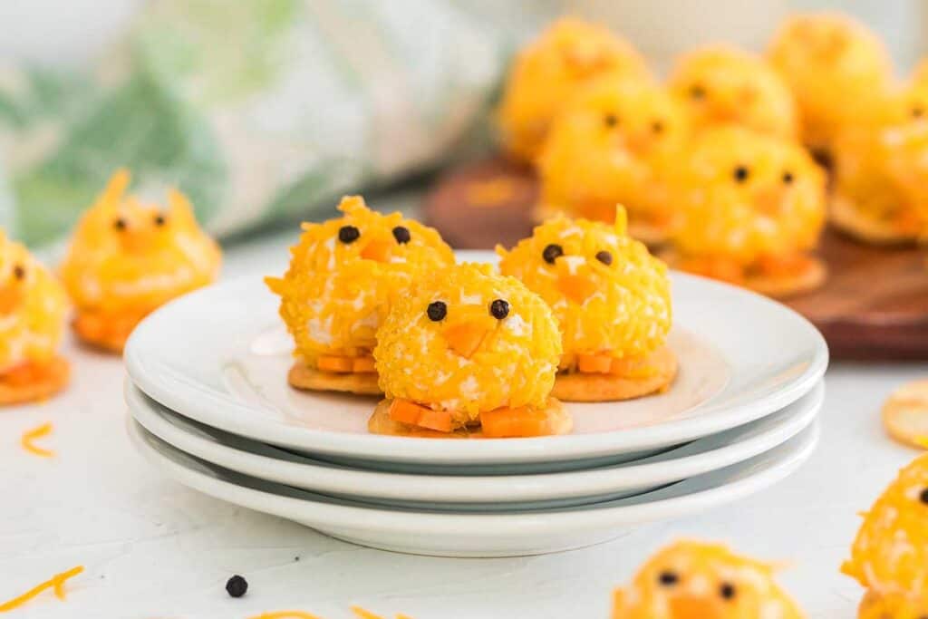 Small, round cheese balls decorated with shredded cheese and black peppercorn eyes are arranged on crackers to resemble chicks, displayed on white plates.