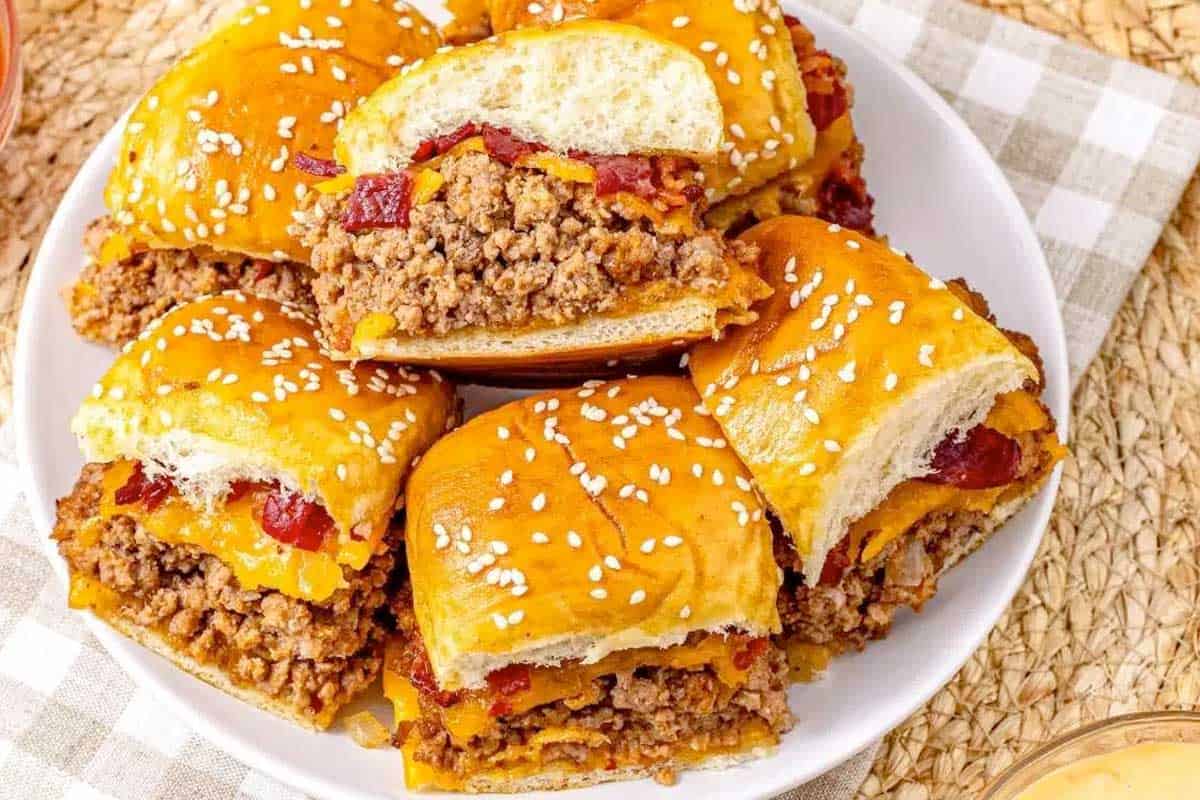 A white plate holds several Bacon Cheeseburger Sliders made with ground beef, crispy bacon, melted cheese, and diced red toppings on sesame seed buns, placed on a checkered cloth and woven surface.