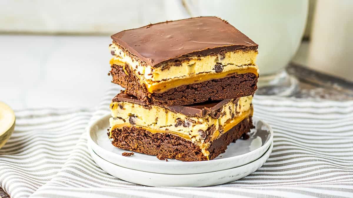Two stacked dessert bars with layers of brownie, caramel, cookie dough, and chocolate topping on a white plate.
