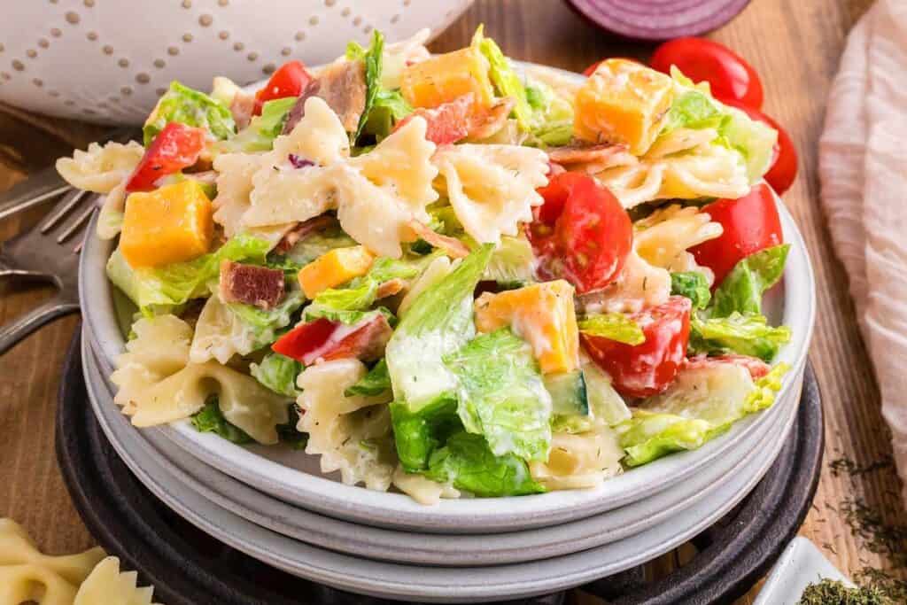A bowl of pasta salad with bowtie pasta, lettuce, cherry tomatoes, diced cheese, bacon bits, and creamy dressing.