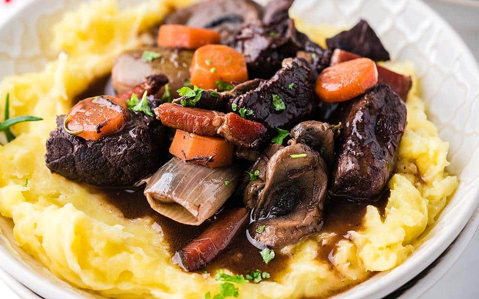 A plate of mashed potatoes topped with beef stew, including carrots, mushrooms, onions, and garnished with herbs.