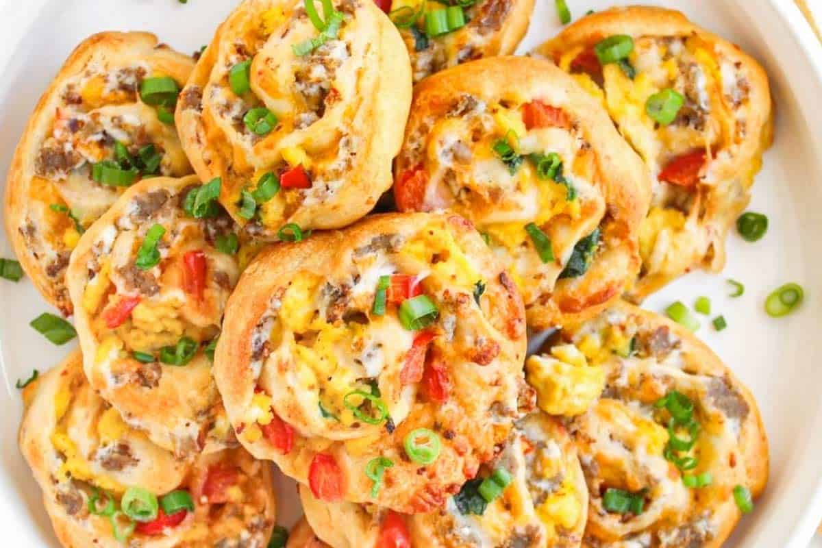 A plate of savory breakfast pinwheels filled with scrambled eggs, sausage, diced red peppers, melted cheese, and topped with chopped green onions.
