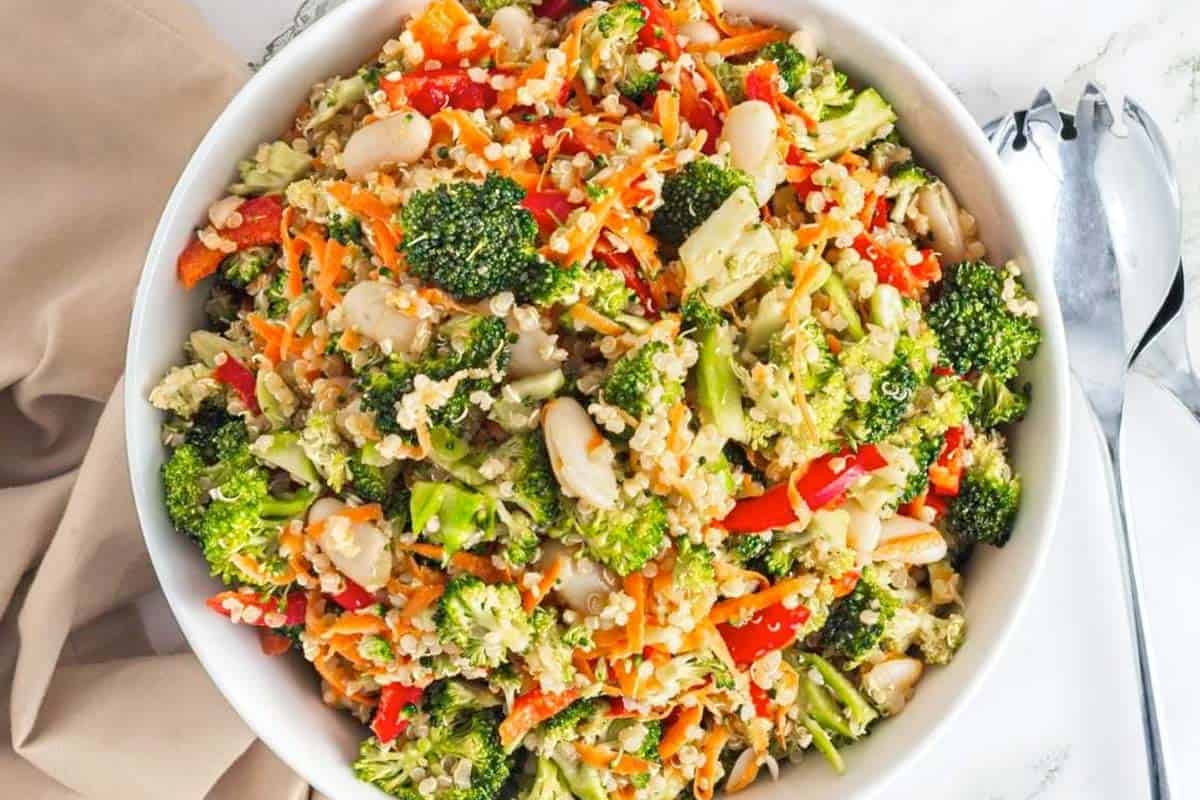 A bowl of mixed salad with broccoli, shredded carrots, red bell peppers, white beans, and quinoa, placed next to a beige napkin and salad serving utensils.