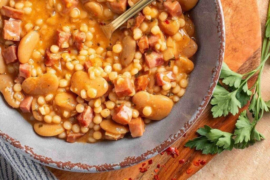 A bowl of bean and pasta soup with diced ham, served on a wooden board next to parsley and a striped cloth.
