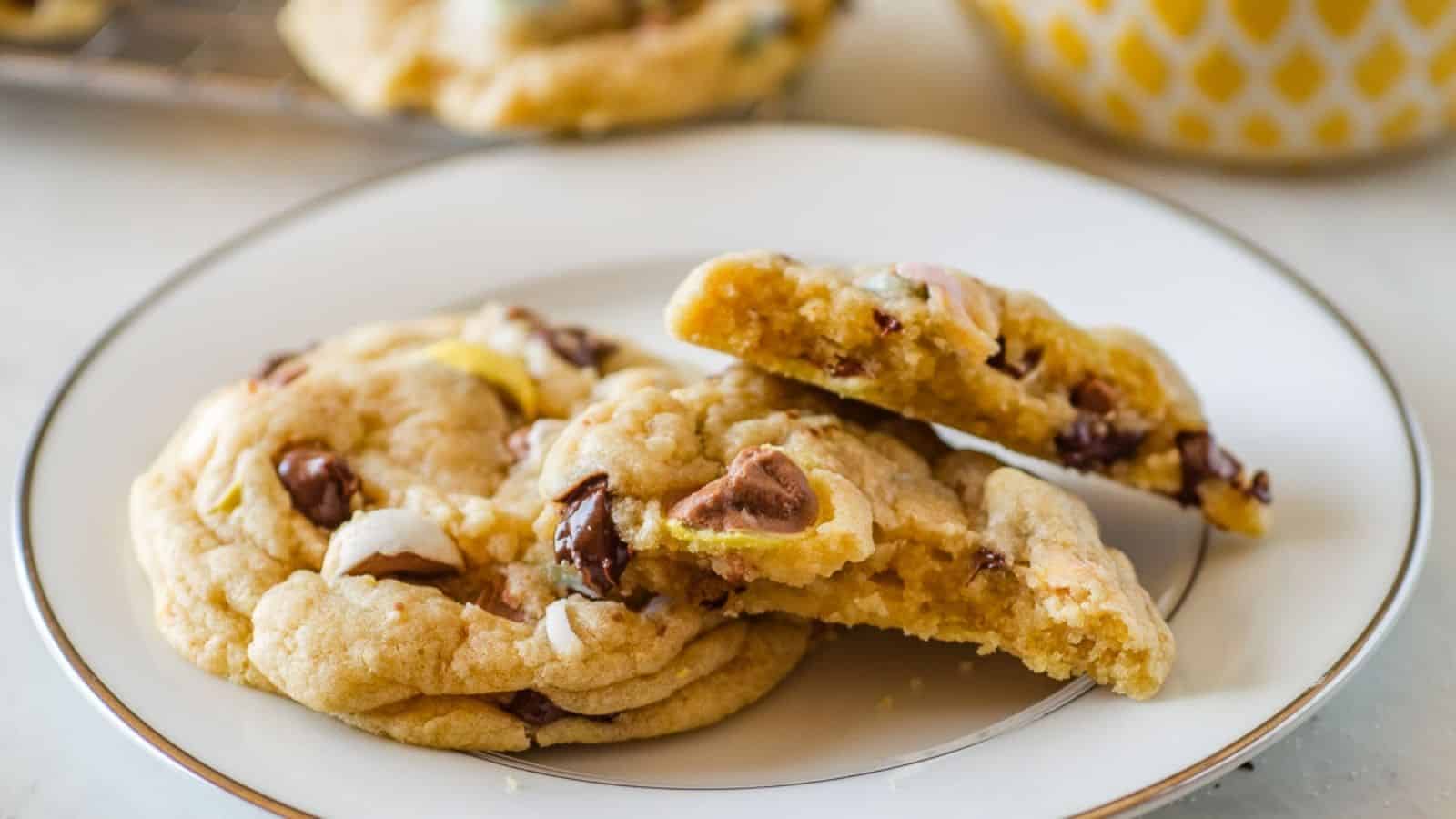 Three chewy cookies with chocolate chunks and perhaps marshmallows on a white plate, with more cookies blurred in the background.