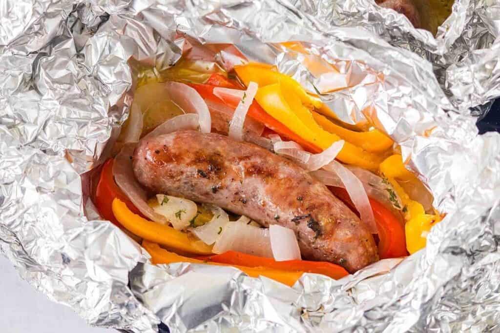A cooked sausage with sliced onions and bell peppers is wrapped in aluminum foil.